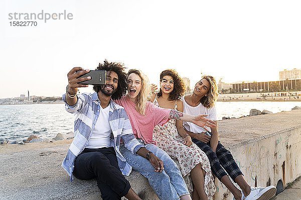 Glückliche Freunde sitzen bei Sonnenuntergang auf der Kaimauer und machen ein Selfie