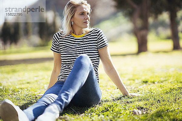 Blond lächelnde Frau  die auf einer Wiese sitzt und seitwärts in einen Park schaut