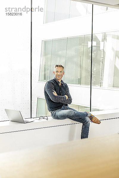 Gelegenheitskaufmann  der auf der Fensterbank eines Bürogebäudes sitzt  mit einem Laptop neben sich