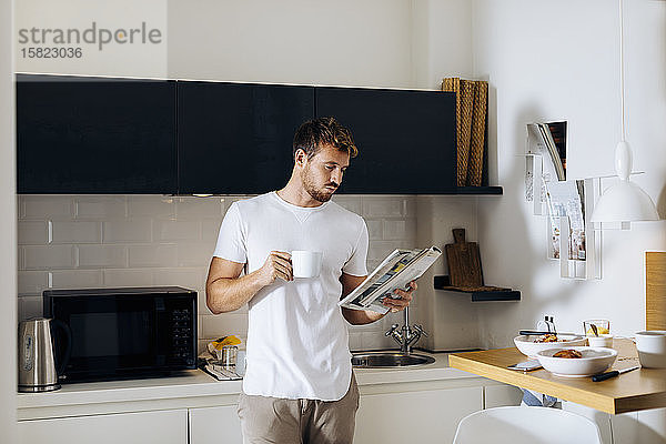 Junger Mann liest eine Zeitschrift und trinkt Kaffee in der Küche