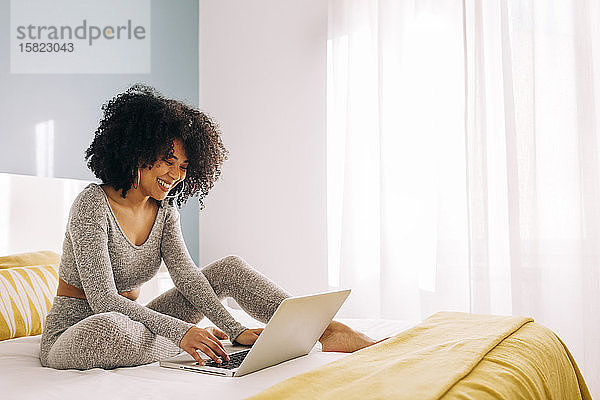 Glückliche junge Frau mit lockigem Haar  die zu Hause einen Laptop auf dem Bett benutzt