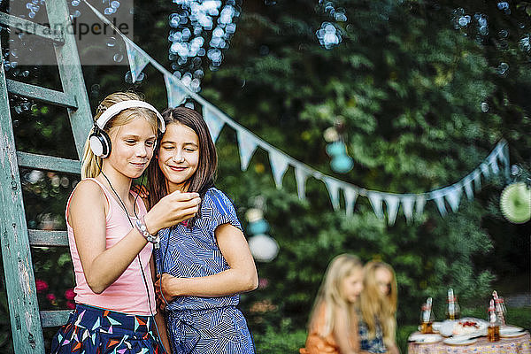 Lächelnde Mädchen mit Kopfhörern und Handy auf einer Geburtstagsparty im Freien