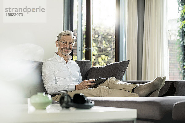 Lächelnder älterer Mann mit grauen Haaren im modern gestalteten Wohnzimmer  der auf einer Couch sitzt und ein Tablett hält