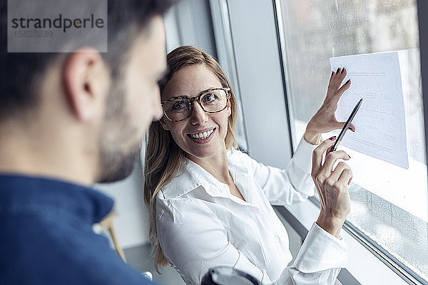 Geschäftsfrau hält ein Blatt Papier am Fenster und zeichnet ein Diagramm  während ein Kollege zusieht