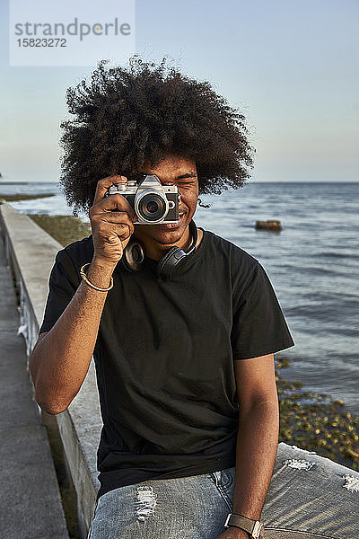 Junger Mann sitzt an der Wand und fotografiert mit seiner Kamera an der Strandpromenade