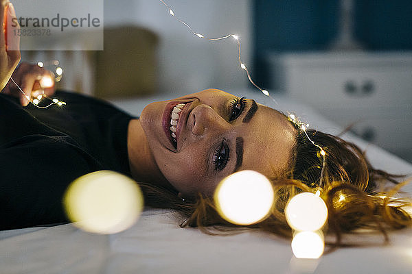 Porträt einer glücklichen jungen Frau im Bett liegend mit Lichterkette
