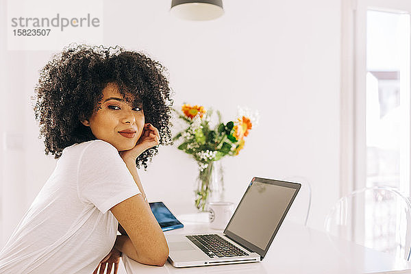 Porträt einer selbstbewussten jungen Frau mit lockigem Haar mit Laptop auf dem Tisch zu Hause