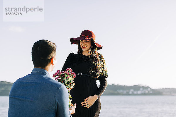 Junger Mann schenkt seiner schwangeren Freundin Blumen an der Uferpromenade