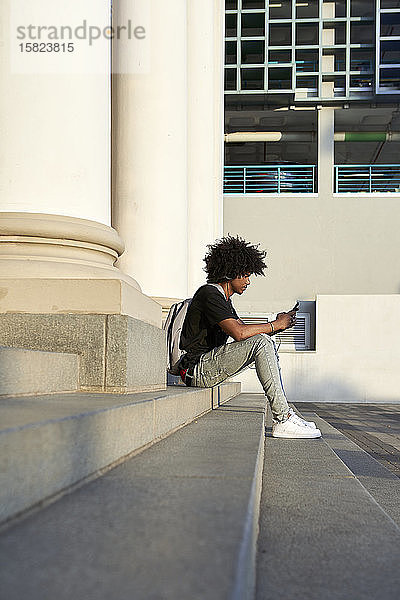 Junger Mann hört Musik  hält sein Smartphone in der Hand  sitzt auf der Treppe