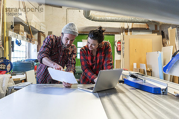 Handwerkerin und Handwerker mit Plan und Laptop diskutieren in ihrer Werkstatt