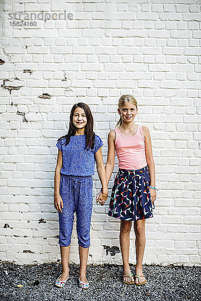 Porträt von zwei lächelnden Mädchen  die Hand in Hand an einer Wand stehen