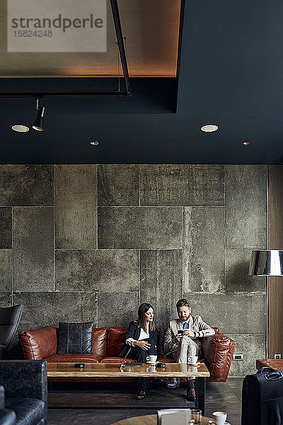 Geschäftsmann und Geschäftsfrau sitzen auf der Couch in der Hotellobby und teilen sich ein Smartphone