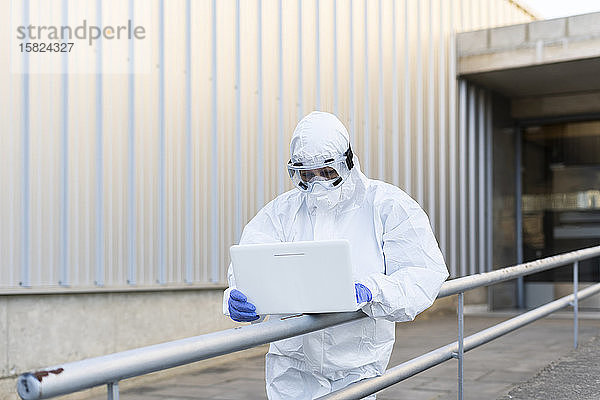 Wissenschaftlerin in Schutzanzug und Maske und mit Laptop vor dem Labor