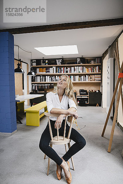 Lächelnde junge Geschäftsfrau sitzt auf einem Stuhl im Loft-Büro und schaut auf