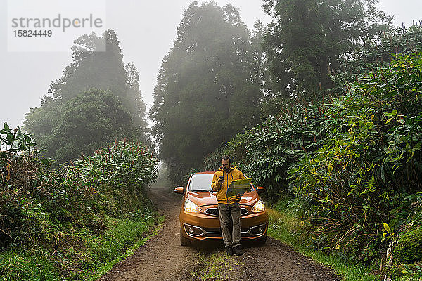 Mann mit Smartphone und Karte vor dem Auto auf Waldstraße  Sao-Miguel-Insel  Azoren  Portugal