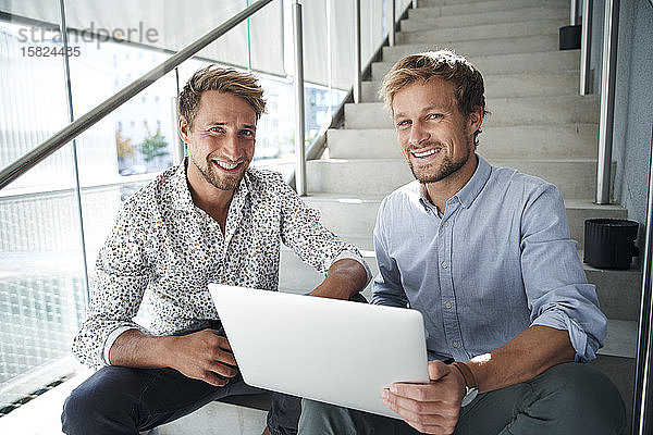 Porträt von zwei lächelnden jungen Geschäftsleuten  die mit Laptop auf einer Treppe sitzen