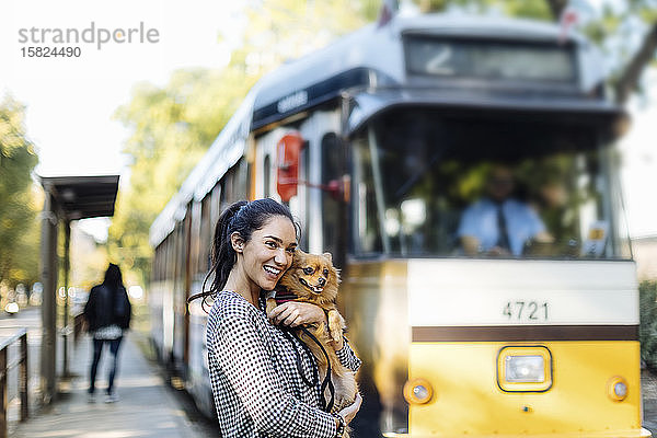Glückliche junge Frau mit Hund an der Straßenbahnhaltestelle in der Stadt