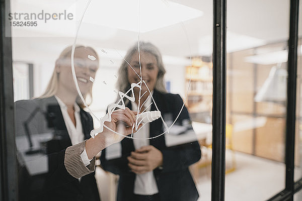 Reife und junge Geschäftsfrau zeichnen im Büro ein Diagramm auf Glasscheibe