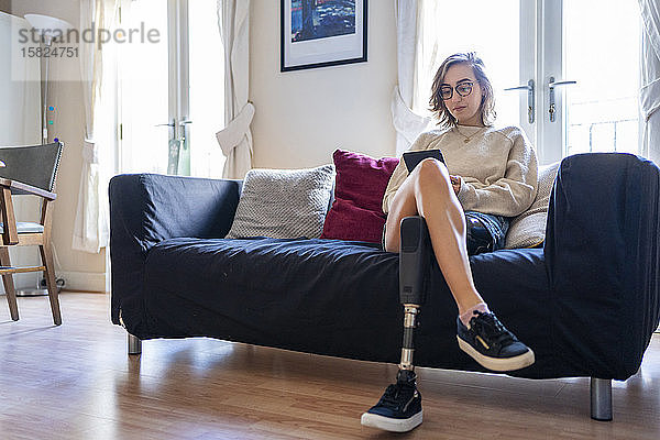 Junge Frau mit Beinprothese sitzt zu Hause auf Couch und liest E-Book