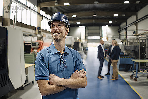 Porträt eines selbstbewussten Arbeiters in einer Fabrik mit Schutzhelm und Geschäftsleuten im Hintergrund