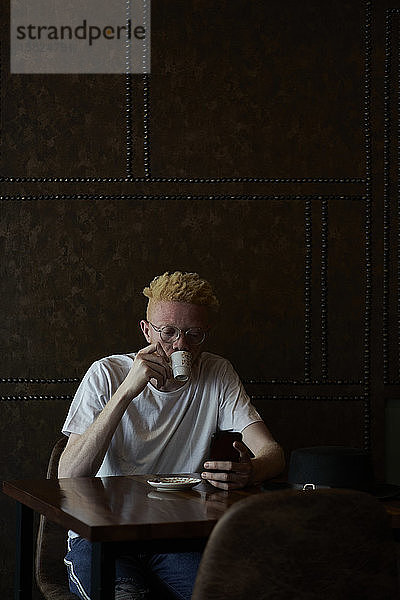 Albinomann mit runder Brille trinkt einen Kaffee und überprüft sein Telefon in einer Cafeteria