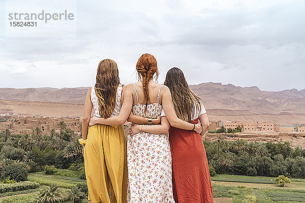 Rückenansicht von drei jungen Frauen  die Arm in Arm stehen und auf die Stadt blicken  Ouarzazate  Marokko