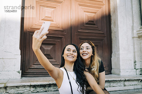 Porträt von zwei glücklichen jungen Frauen  die sich mit einem Smartphone selbstständig machen  Lissabon  Portugal