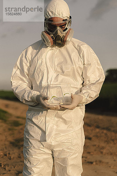 Mann mit Schutzanzug und Maske auf dem Land mit Keimling in Glasvitrine