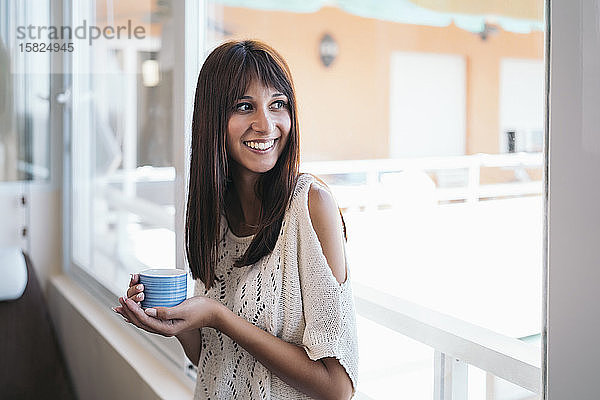 Porträt einer lächelnden jungen Frau mit Kaffeetasse nahe am Fenster zu Hause