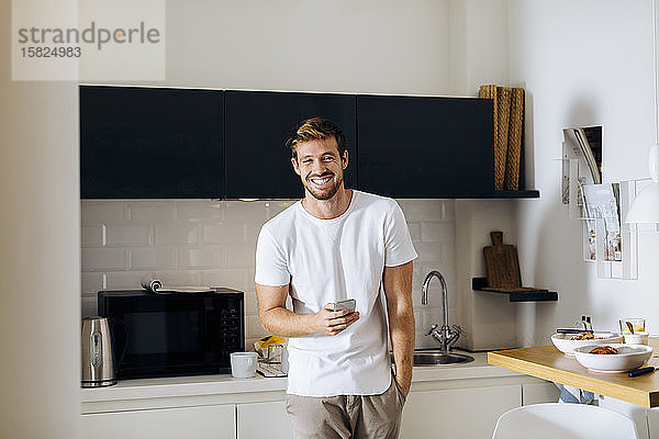 Porträt eines glücklichen jungen Mannes mit Handy in der Küche