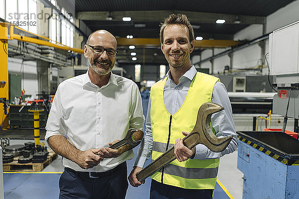 Porträt eines Geschäftsmannes und eines Mannes in reflektierender Weste mit großen Schraubenschlüsseln in einer Fabrik