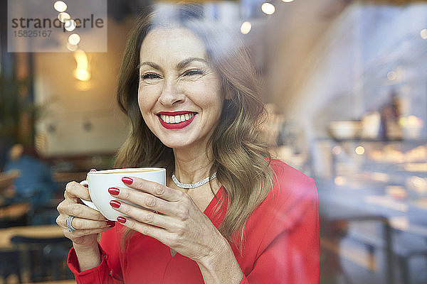 Porträt einer lachenden  reifen Frau mit einer Tasse Kaffee hinter einer Fensterscheibe