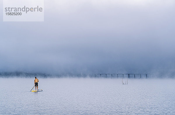 Mann steht beim Paddel-Surfen auf einem See im Nebel auf
