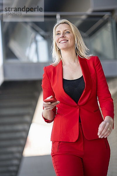 Blond lächelnde Geschäftsfrau im roten Anzug mit Smartphone in der Hand