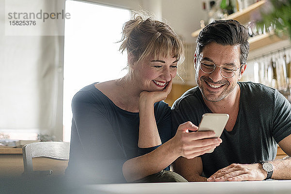 Glückliches Paar sitzt am Tisch in der Küche und benutzt ein Smartphone