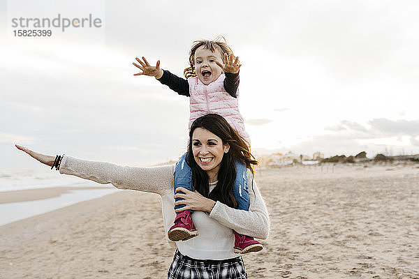 Glückliche Mutter trägt ihre Tochter bei Sonnenuntergang am Strand auf den Schultern