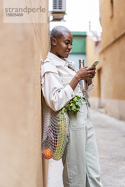 An die Wand gelehnte Frau mit Thermoskanne und Lebensmitteln per Smartphone