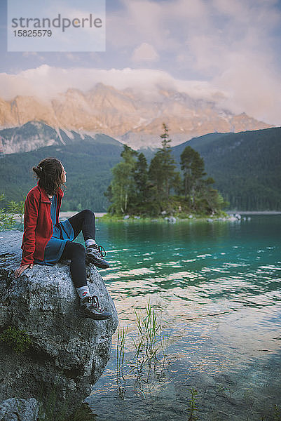 Deutschland  Bayern  Eibsee  Junge Frau sitzt auf einem Felsen und schaut auf die Aussicht am Eibsee in den bayerischen Alpen
