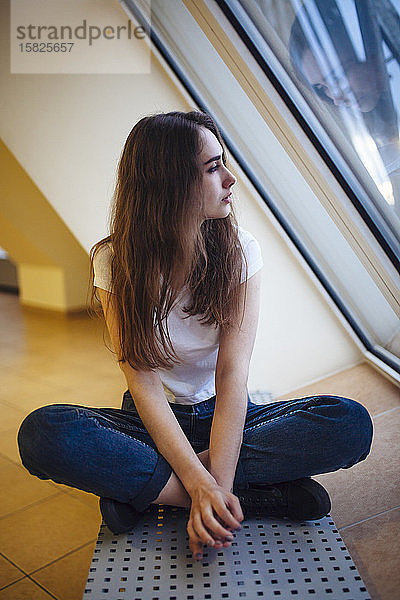 Junge Frau sitzt auf dem Boden und schaut durch das Fenster