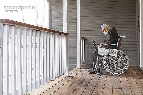 Älterer Mann im Rollstuhl trägt Schutzmaske  um die Übertragung des Coronavirus zu verhindern  und benutzt einen Laptop auf der Veranda
