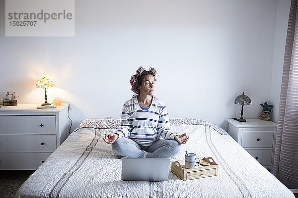 Frau mit Lockenwicklern meditiert auf einem Bett vor einem Laptop