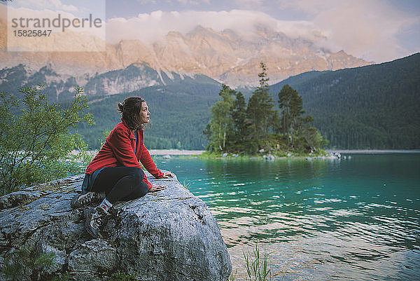 Deutschland  Bayern  Eibsee  Junge Frau sitzt auf einem Felsen und blickt auf den Eibsee in den Bayerischen Alpen