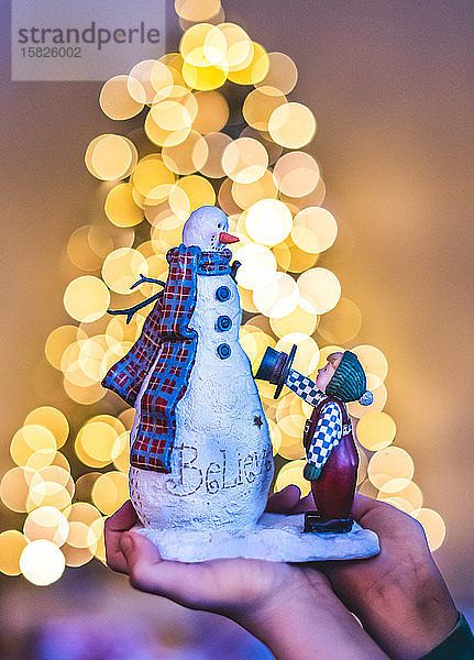 Nahaufnahme der Hände  die eine Schneemann-Dekoration mit Lichtern im Hintergrund halten