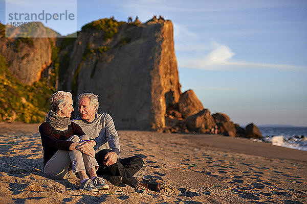 Älteres Ehepaar unterhält sich bei Sonnenuntergang am Strand im Sand sitzend