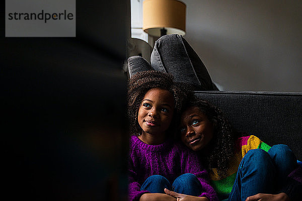 Nachdenklich lächelnde Schwestern sitzen zu Hause gegen das Sofa im Wohnzimmer