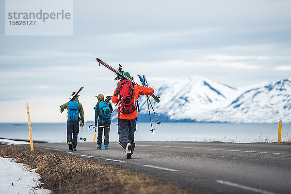 Drei Personen mit Skiern gehen in Island eine Straße entlang
