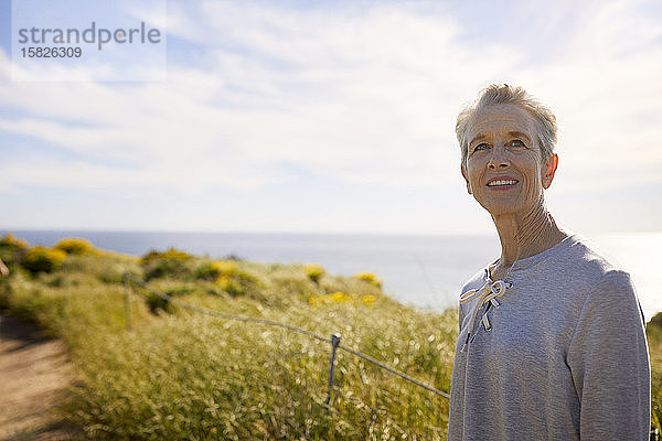 Nachdenkliche ältere Touristin  die im Sommer auf einer Klippe am Meer gegen den Himmel steht