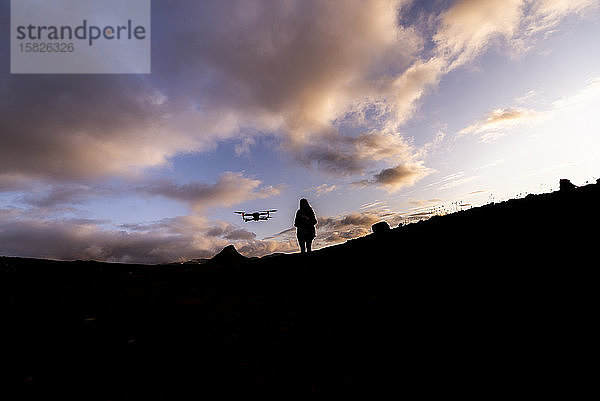 Silhouette einer Frau  die bei Sonnenuntergang eine Drohne kontrolliert und steuert