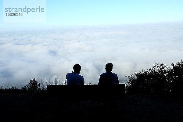 zwei Personen  die auf einer Bank auf einem Berggipfel sitzen und Wolken überblicken