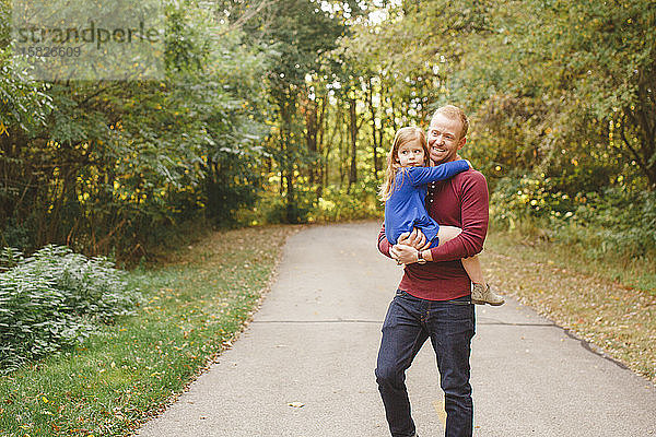 Ein lächelnder Vater hält eine kleine Tochter auf einem von Bäumen gesäumten Weg in den Armen